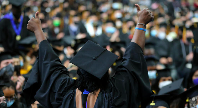 Person wearing graduation cap and gown facing away from the camera towards a crowd with arms raised and thumbs up