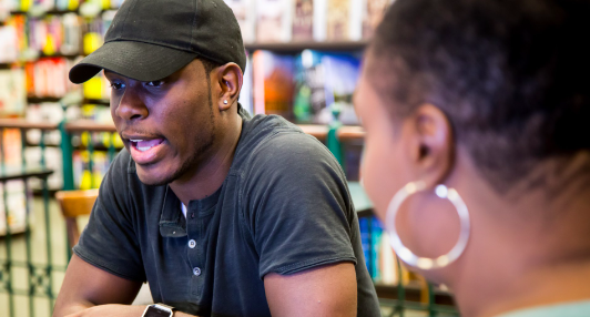 Black man with cap on talking to person with silver earrings