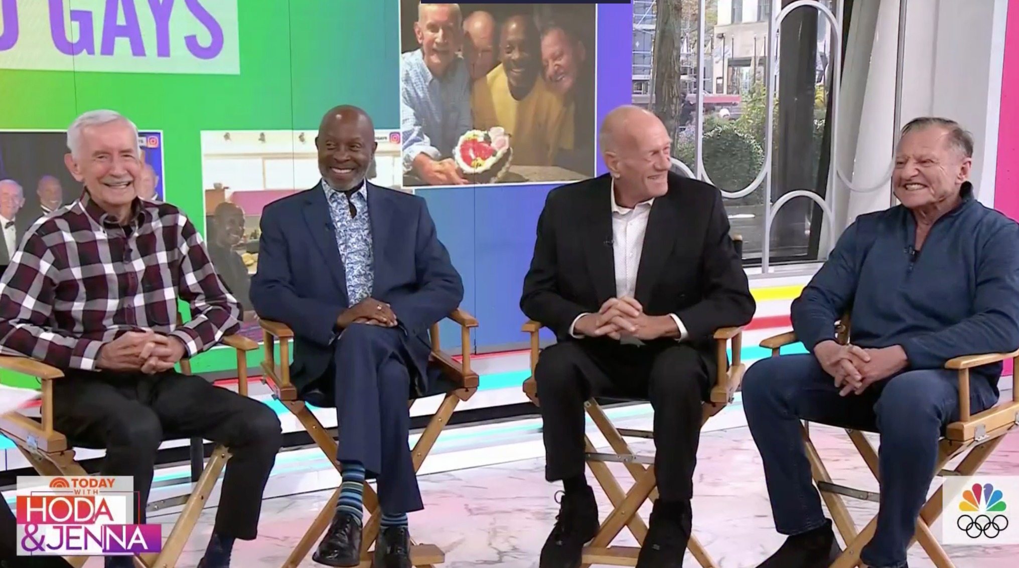 Four elderly gay men sit on a studio set smiling 